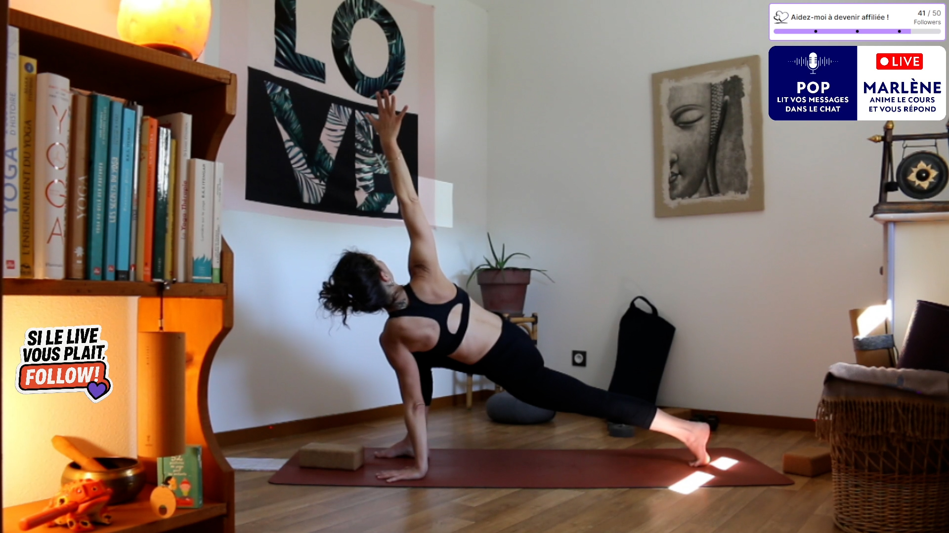 Personne pratiquant une pose de yoga à l'intérieur lors d'un cours en direct, avec des étagères et des œuvres d'art murales en arrière-plan.